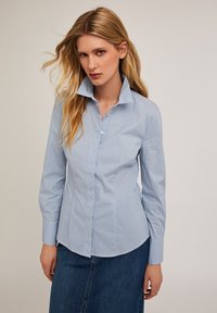 Camisa de botón azul claro hecha de algodón a rayas. Cuenta con un cuello alto, mangas largas y un corte entallado. Textura suave.