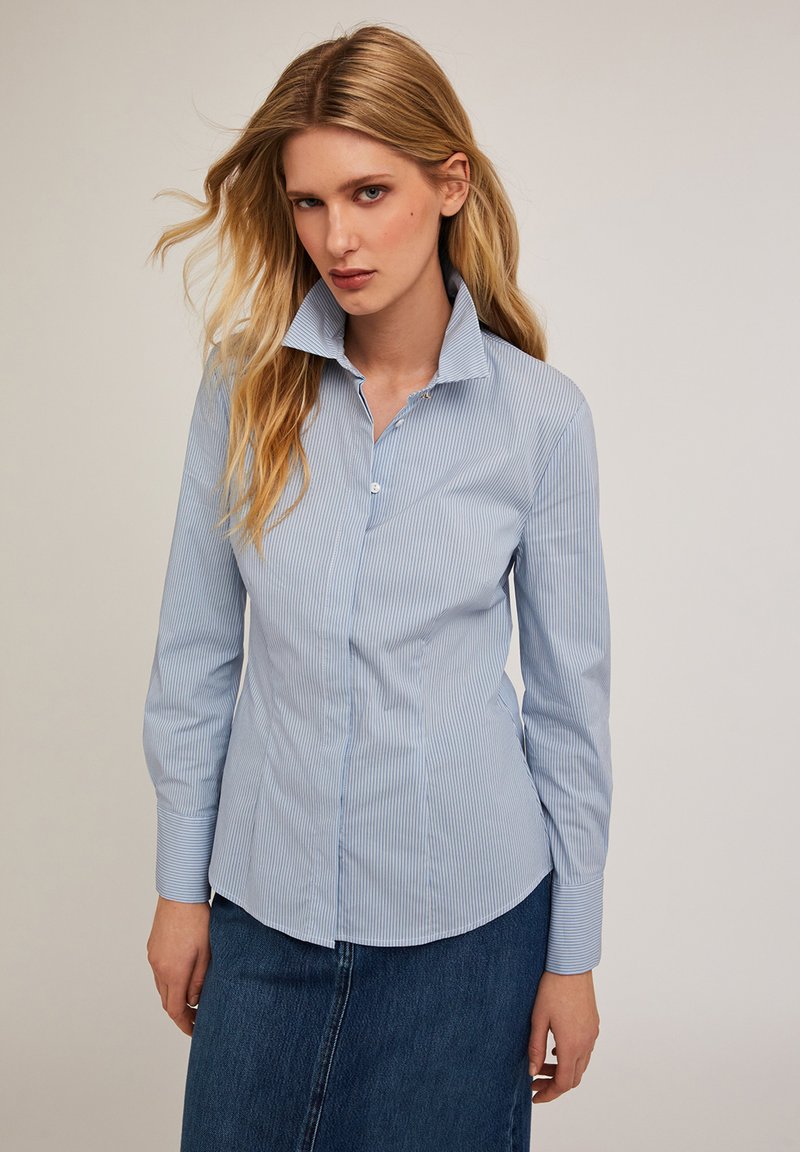Camisa de botón azul claro hecha de algodón a rayas. Cuenta con un cuello alto, mangas largas y un corte entallado. Textura suave.