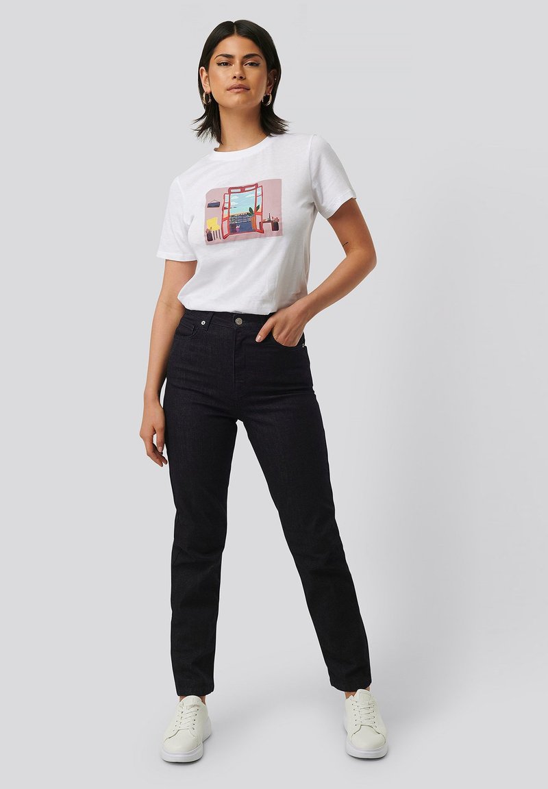 Kvinna som bär en vit t-shirt med grafisk tryck, svarta högmidjade jeans och vita sneakers, stående med ena handen i fickan mot en enfärgad bakgrund.