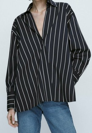 Personne portant une chemise noire ample à manches longues avec des rayures verticales blanches et un jean bleu sur un fond clair uni.