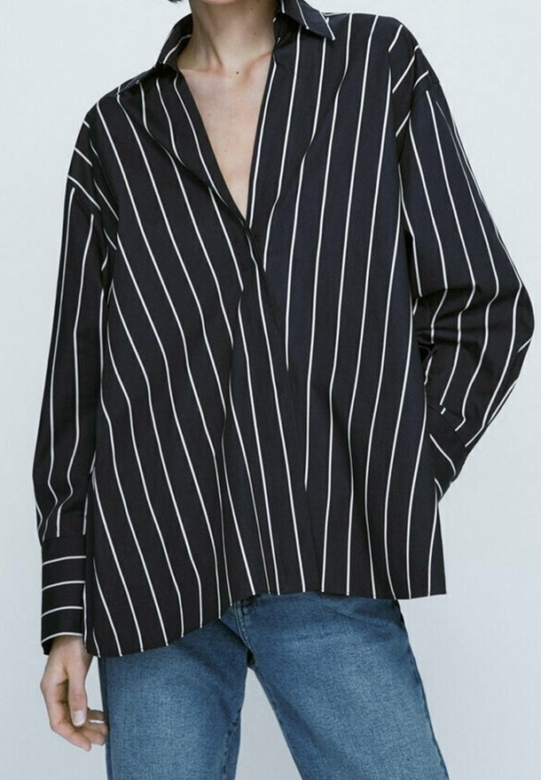 Personne portant une chemise noire ample à manches longues avec des rayures verticales blanches et un jean bleu sur un fond clair uni.