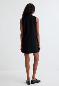 Schwarz ärmelloses Kleid mit hohem Kragen, gerader Silhouette, Reißverschluss hinten, kombiniert mit schwarzen flachen Schuhen und strukturierten Akzenten.