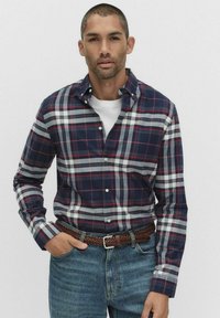 Koszulka w granatowo-białą krateczkę z czerwonymi akcentami, z długimi rękawami, kołnierzyk zapinany na guziki oraz biała koszulka pod spodem. Noszona z niebieskimi dżinsami i brązowym plecionym paskiem.