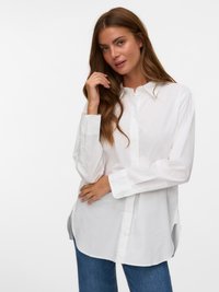 Young woman with long brown hair wearing a loose white button-up shirt and blue jeans, posing with one hand near her face.