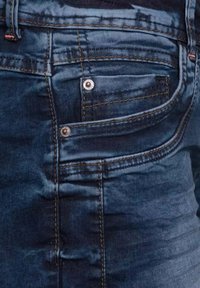 Farkkutakki tummansinisellä, hieman haalistuneella pinnalla. Varustettu pienellä taskulla, metallinapeilla ja erottuvalla keltaisella ompelulla.