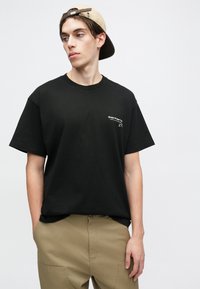 Tricou din bumbac negru, cu guler rotund și mâneci scurte. Prezintă un mic logo alb imprimat pe pieptul stâng. Este purtat cu pantaloni bej.