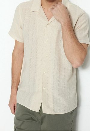 Homme portant une chemise beige à manches courtes à boutons avec des rayures verticales texturées et un pantalon vert olive, bras levé jusqu'à la poitrine.