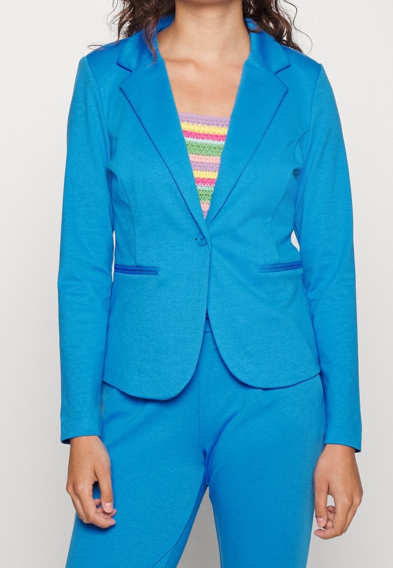 Femme portant un blazer bleu vif et un pantalon assorti, avec un haut à rayures colorées en dessous, sur un fond uni.