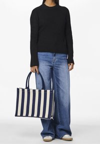 Pull en tricot noir, large jean denim bleu clair, tenant un sac cabas rayé bleu marine et crème avec deux poignées. Style décontracté, coupe ample.