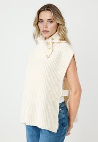 Gilet in maglia crema con colletto alto, spacchi laterali, dettagli con bottoni dorati e motivo a coste testurizzato. Indossato sopra una maglietta aderente e jeans.