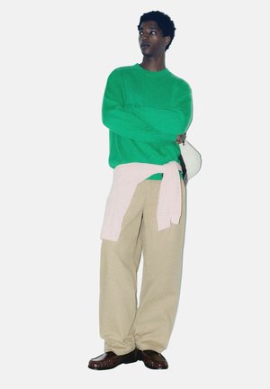 Jeune personne debout, les bras croisés, portant un pull vert, un pantalon large beige, un pull rose noué à la taille, et tenant un chapeau blanc.
