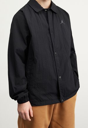 Osoba ubrana w czarną kurtkę zapinaną na guziki z elastycznymi mankietami i małym logo na piersi, w połączeniu z jasnobrązowymi spodniami.