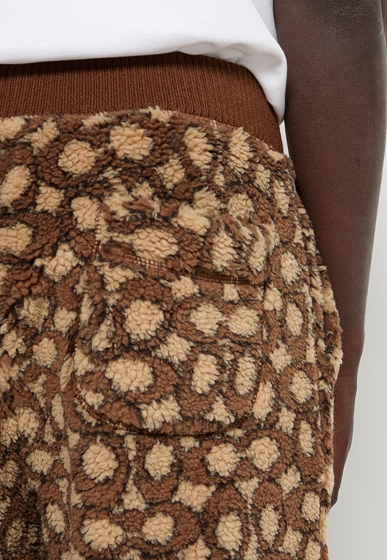 Brązowe i beżowe spodnie polarowe w leopardzi wzór z kieszonką na boku, noszone przez osobę w białej koszuli.
