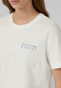 White cotton t-shirt featuring a crew neck and short sleeves. Printed text in blue reads "ENJOY EVERY MOMENT" on the chest.