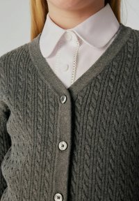 Grau gestrickte Strickjacke mit einem Zopfmuster, V-Ausschnitt, sechs Knopfverschlüssen und über einem weißen Hemd mit Kragen getragen.