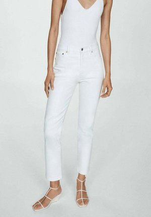 Frau, die eng anliegende weiße Jeans, ein ärmelloses weißes Top und weiße Keilsandalen mit Riemchen trägt, steht vor einem einfachen hellen Hintergrund.