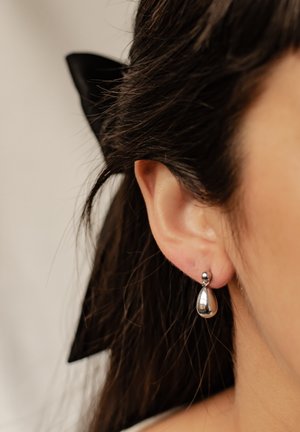 Silver teardrop earring with a polished surface, attached by a small spherical stud, worn on an ear with long dark hair and a black ribbon.