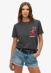 Camiseta de algodón gris con diseño bordado colorido que incluye alas, rosas y texto. Combinada con pantalones cortos de mezclilla azul claro y un cinturón negro.