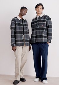 Zwei Modelle tragen karierte Flanellhemden in Schwarz, Grau und Blau mit einem weißen Kragen. Ein Modell kombiniert es mit beigefarbenen Cargo-Hosen, das andere trägt eine marineblaue Hose.