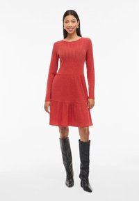 Červené žebrované šaty s dlouhými rukávy, kulatým výstřihem a rozšířenou sukní. Nosit s vysokými černými botami, které mají texturovaný povrch.