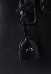 Czarna skórzana torba z prostokątną metką z wytłoczonym złotym logo "A/X" oraz detalem w postaci plecionej pętli, z gładką fakturą.