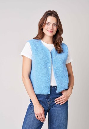 Vrouw met bruin haar die een lichtblauw gestructureerd fleecevest draagt over een wit shirt en blauwe spijkerbroek, staand met één hand op haar heup.