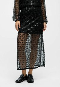Schwarzes Paillettenkleid mit durchsichtiger Überlage, ausgestattet mit langen Ärmeln und einem schmalen Schlitz bis zum Oberschenkel. Getragen mit schwarzen Slippern, die eine kontrastierende Textur zeigen.