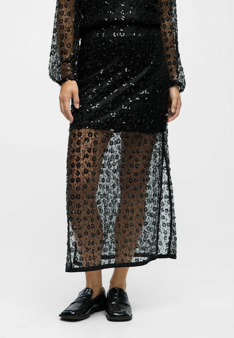 Schwarzes Paillettenkleid mit durchsichtiger Überlage, ausgestattet mit langen Ärmeln und einem schmalen Schlitz bis zum Oberschenkel. Getragen mit schwarzen Slippern, die eine kontrastierende Textur zeigen.