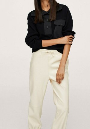 Femme portant un haut noir à manches longues boutonné et un pantalon crème à cordon de serrage, debout devant un fond uni.