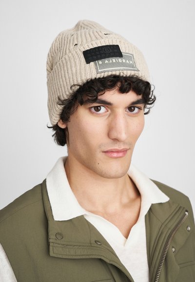 Gorro de malha bege com textura canelada, apresentando um patch retangular preto com texto branco e um design de calendário, usado com um colete oliva.