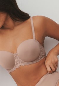 Donna che indossa un reggiseno beige imbottito con dettagli in pizzo, che sistema la biancheria intima coordinata su uno sfondo chiaro e uniforme.
