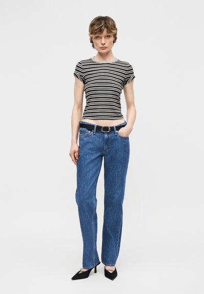 Νεαρή γυναίκα στέκεται φορώντας κοντομάνικη μπλούζα με ασπρόμαυρες ρίγες, μπλε ψηλόμεσο τζιν με μαύρη ζώνη και μαύρες μυτερές γόβες.