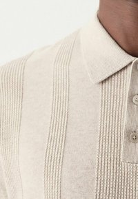 Polo en tricot beige avec un design à rayures texturées, un col et trois boutons au niveau de l'encolure. Tissu doux avec une coupe décontractée.