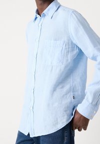 Camicia azzurro chiaro in lino, con taschino sul petto, colletto a punta e maniche lunghe con polsini abbottonati.