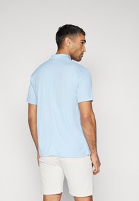 Polo de cor azul claro feito de tecido suave, com manga curta, colarinho e detalhe na costura das costas. Usado com calções brancos.