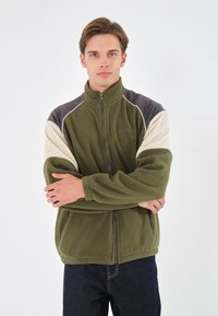 Giacca in fleece verde scuro, con colletto alto e zip frontale. Maniche a contrasto in crema e grigio scuro. Texture morbida, vestibilità casual.