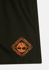 Olivovo zelená textília obsahuje jasne oranžové logo Timberland: stylizovaný strom v tvare diamantu, obklopený textom a okrajom.