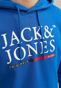 Modrá mikina s kapucí se bílým nápisem "JACK & JONES", černým a červeným pruhem dole a potiskem "ORIGINALS ©1990" na přední straně.