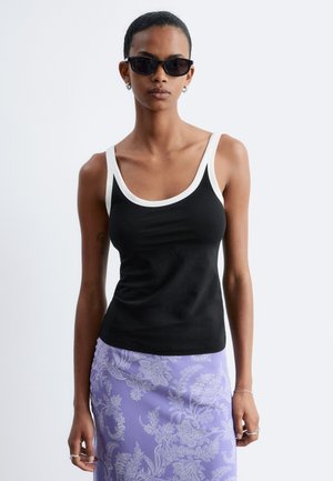 Femme portant des lunettes de soleil noires, un débardeur noir avec bordure blanche et une jupe violette à motif cachemire sur un fond uni.