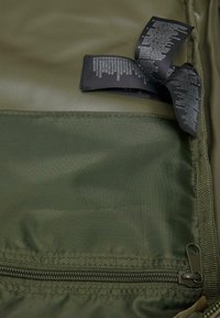 Green textile bag featuring a zippered pocket at the bottom and care tags in black textile showing care instructions. Smooth, matte texture.