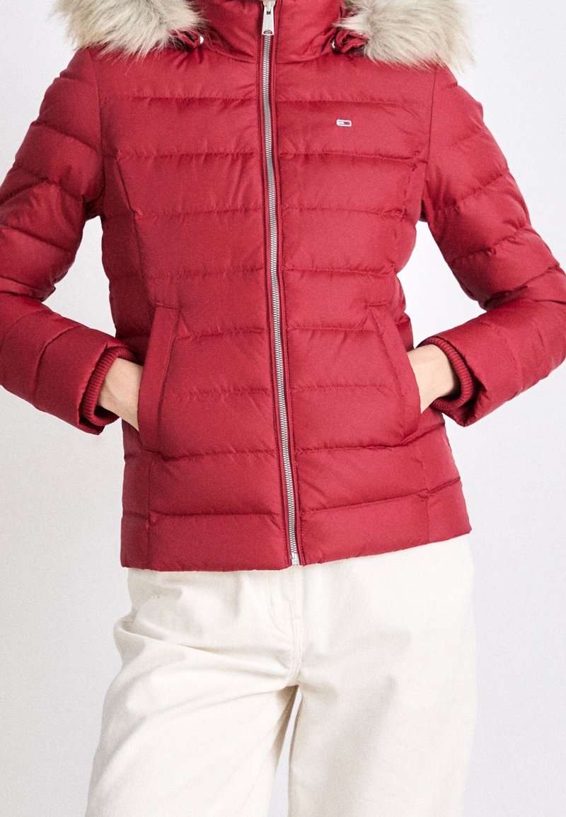 Veste rouge matelassée avec capuche bordée de fourrure, fermeture éclair à l'avant et mains dans les poches, portée avec un pantalon de couleur claire.