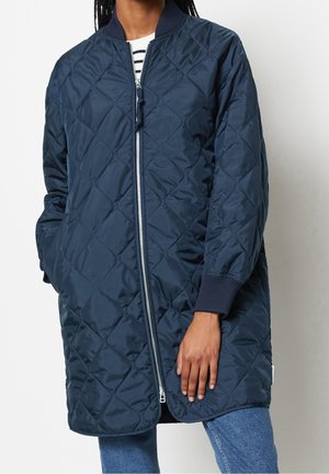Personne portant une veste bomber matelassée bleu marine avec des poignets côtelés, une fermeture éclair sur le devant, par-dessus une chemise rayée et un jean bleu.