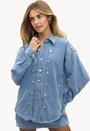 Femme aux longs cheveux blonds portant une veste en jean bleue oversize avec des boutons argentés et une jupe courte assortie, regardant sur le côté.