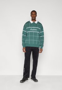 Πράσινο πουλόβερ με σχέδια και λευκές ρίγες, με το κείμενο "Malbon"· συνδυασμένο με μαύρο παντελόνι από κορδόνι με πλαϊνή κεντητική και μαύρα παπούτσια.