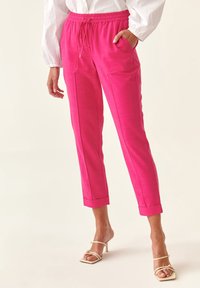 Svetle růžové, lehké kalhoty s páskem na stažení v pase, předními kapsami, slim fit a ohrnutými manžetami. Nošeny s páskovými, neutrálními podpatky.