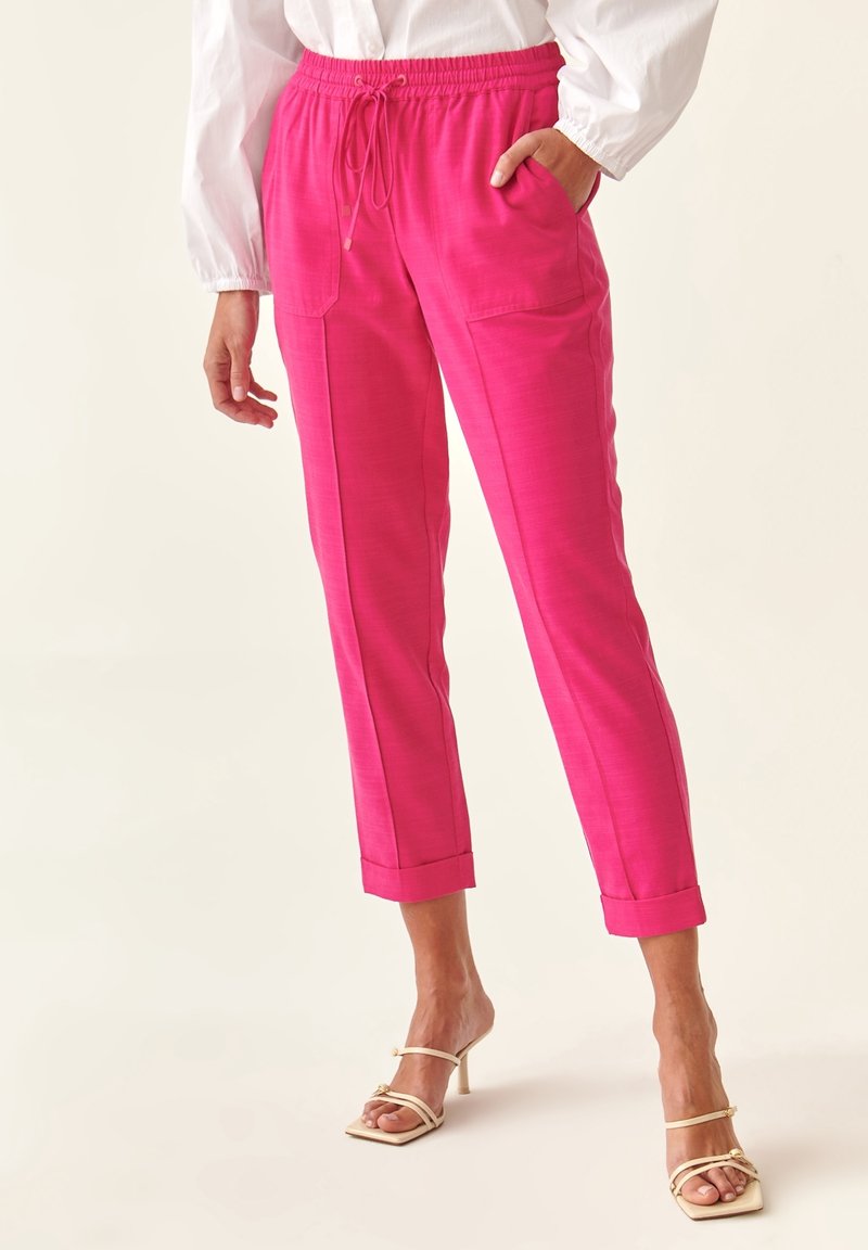 Svetle růžové, lehké kalhoty s páskem na stažení v pase, předními kapsami, slim fit a ohrnutými manžetami. Nošeny s páskovými, neutrálními podpatky.