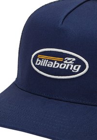 Casquette bleu marine à visière plate, ornée d'un logo Billabong brodé en blanc et jaune dans un design ovale. Détails en tissu texturé visibles.