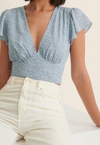 Haut court à fleurs bleu clair avec manches courtes à volants, encolure en V profonde et fronces sous la poitrine, associé à un jean blanc taille haute.