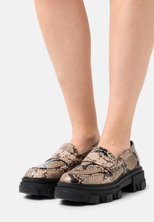 Mocasines con patrón de piel de serpiente en beige y negro con suelas negras gruesas, que presentan una textura suave y un diseño de punta redondeada.