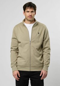 Beige zip-up sweatshirt lavet af blødt materiale, med en standkrave og et lille marineblåt logo på venstre bryst, parret med mørke jeans.